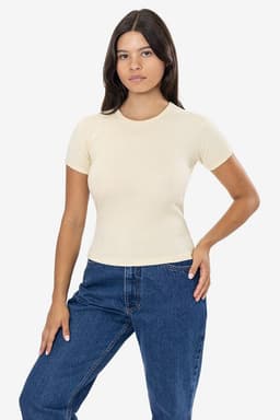 A woman wearing a light beige short-sleeved baby rib tee and blue jeans, standing confidently. The tee has a logo.