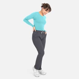 A woman wearing a turquoise long-sleeve tee and grey pants, smiling and looking down at her outfit.