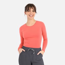 A woman in a coral long-sleeve top, smiling while wearing grey pants. The top features a logo.