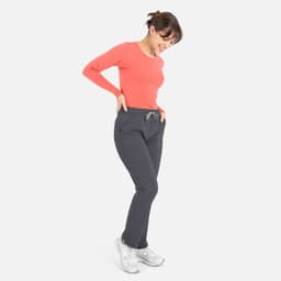 A woman wears a coral long-sleeve underscrub tee with grey pants, smiling and posing. The outfit features pockets.