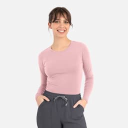 A woman wears a pink long-sleeve underscrub tee paired with grey pants and has her hands in her pockets.