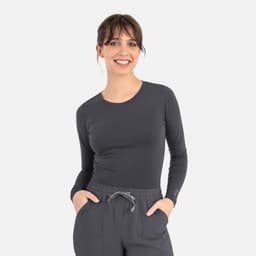 A long-sleeve women's underscrub tee in grey, worn by a model with hands in pockets, featuring a logo.