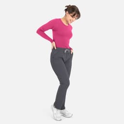 A woman wears a pink long-sleeve underscrub tee with grey pants and white sneakers, smiling while looking down.