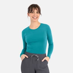A long-sleeve teal underscrub tee paired with grey trousers, modelled by a smiling woman with her hands in pockets.