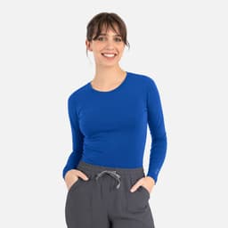 A woman wearing a long-sleeve, bright blue underscrub tee with a fitted style and grey pants. The tee has a logo.