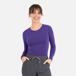 A purple long-sleeve underscrub tee worn by a smiling woman, paired with grey pants. The tee features a logo.