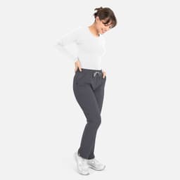 A woman wearing a white long-sleeve underscrub tee and grey pants, smiling with hands in pockets.