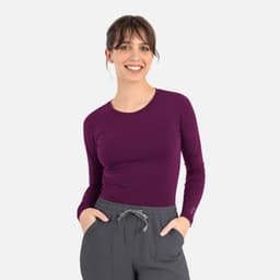 A woman smiles while wearing a long-sleeve underscrub tee in deep purple, paired with grey pants.