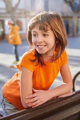 A girl leans on a railing, wearing an orange basic T-shirt, smiling in a sunlit outdoor setting.
