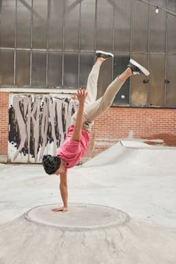 A person performing a handstand in a pink T-shirt and beige pants at a skate park with industrial background.