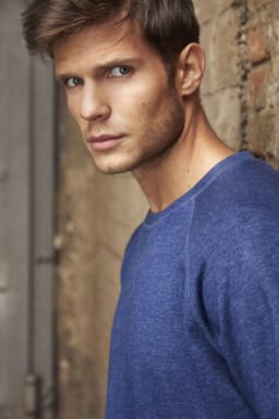 A blue men's sweater is worn by a model standing against a textured wall. The garment has a relaxed fit and casual style.