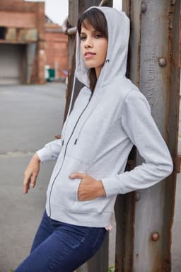A grey ladies hoodie with a zip and pockets, worn by a model against a metal structure.