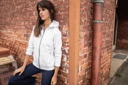 A zip hoodie in light grey displayed on a model against a brick wall, featuring a hood and zip closure.
