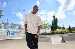 An oversized cream T-shirt worn by a model, set against a sunny outdoor skatepark backdrop.