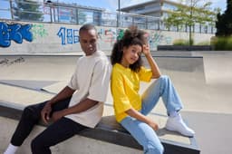 Two models seated by a skate park, wearing oversized t-shirts; one in beige and the other in yellow, with casual jeans and sneakers.