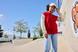 A varsity jacket in red and beige is worn by a person standing in a skate park, with a cap and blue jeans.