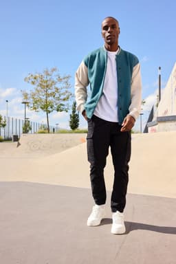 A varsity jacket in teal and cream, worn over a white shirt, paired with black cargo pants and white sneakers.