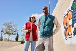 A red and teal varsity jacket worn by two individuals outdoors, with a graffiti background and blue sky above.