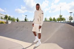 An oversized hooded jacket in beige is worn with matching shorts, set in a sunny skatepark. The model stands on a ramp.