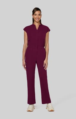 A woman stands in a burgundy jumpsuit with cap sleeves, featuring a front zip and pocket details.