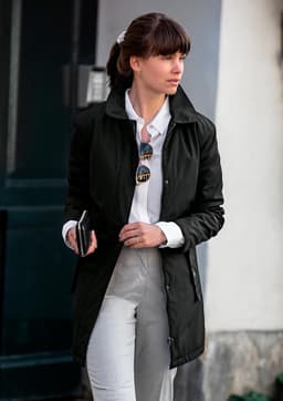 A stylish black women's jacket is featured, worn over a white shirt and grey pants, accessorised with sunglasses.