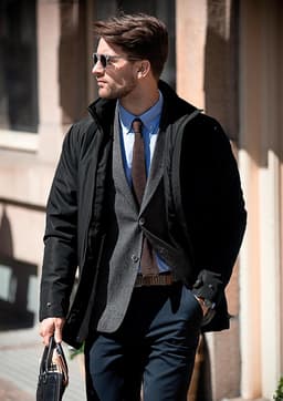 A stylish black men's jacket worn over a grey blazer with a light blue shirt, paired with sunglasses and trousers.