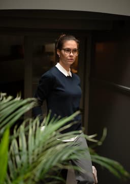 A woman wears a navy knitted jumper paired with a collared shirt, walking past a green plant indoors.