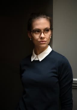 A model in a navy knitted jumper with a white collar, wearing glasses and standing against a dark background.