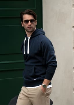 A navy hoodie with a drawstring and front pocket, worn by a man with sunglasses, standing against a green door.