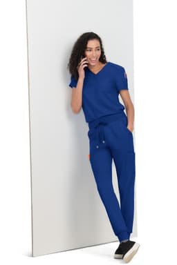 A woman wearing a blue scrub set, leaning against a white panel, with curly hair and a casual pose.