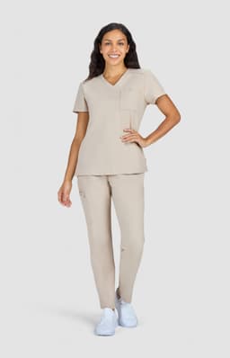 A woman models a beige scrub set with a pocketed top and matching pants, complemented by white shoes.