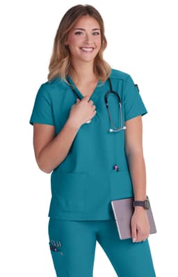 A medical scrub top in teal, featuring a round neckline, pockets, and a stethoscope. The model is smiling and holding a tablet.