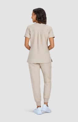 A woman wears a beige medical uniform with a pleated back and matching trousers, paired with white shoes.