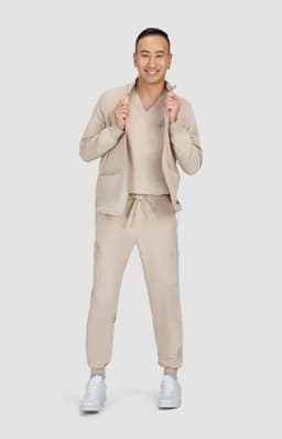 A man is wearing a beige jacket with a matching top and trousers, standing in a neutral setting. The outfit features a logo.