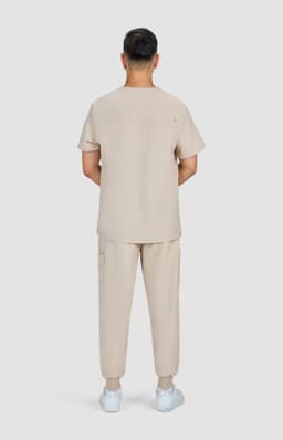 A man stands with his back to the camera, wearing a beige medical uniform consisting of a short-sleeve top and pants.