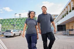 A man and a woman wearing grey work polo shirts, walking together outside a building. The setting features greenery in the background.