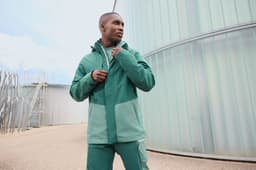 A model wearing a green hardshell rainjacket, featuring contrasting shades and a logo, set in an outdoor industrial environment.