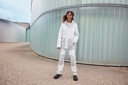 A model wearing a white and grey hardshell rainjacket with a hood, paired with matching pants and black shoes.