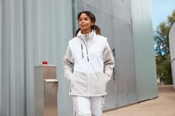 A woman wearing a grey and white softshell vest, walking outdoors near a metal building. The vest has a logo.