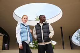 A blue ladies winter vest and a black men's vest are worn by two models outdoors under a circular structure.