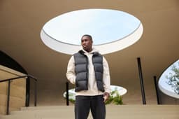A man wears a black puffer vest over a beige hoodie, posing outdoors under an architectural structure.