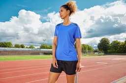 A blue ladies running t-shirt with short sleeves, displayed on a model near a running track. It has a logo.
