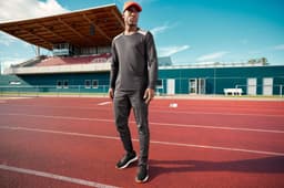 A long-sleeve men's running shirt in grey, paired with black pants and a red cap, on a track field.