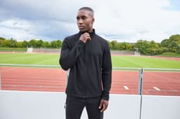 A man wearing a black structured fleece jacket stands outdoors, with a sports track visible in the background.