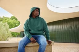 A unisex oversized sweat hoody in green, worn by a person sitting on a ledge, with a modern architectural background.