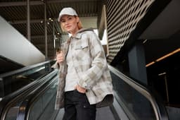 A check shirt jacket in grey tones, featuring a hood, worn by a model in a modern indoor setting.