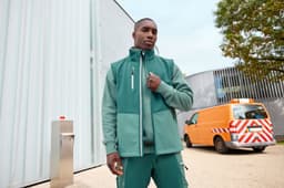 A model wearing a green softshell vest stands outdoors, paired with matching green workwear. A vehicle is visible in the background.