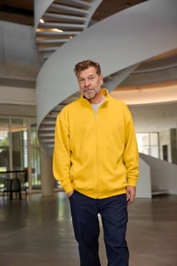 A man wearing a yellow sweat jacket walks through a bright, modern space with a spiral staircase in the background.