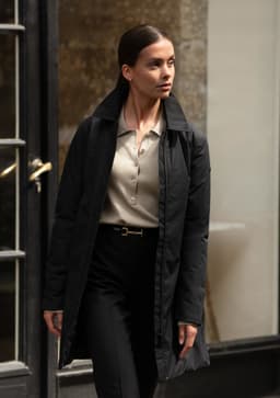 A woman in a black jacket walks outside, paired with a beige top and black trousers. The jacket has a logo.