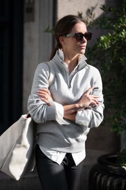 A grey unisex sweatshirt with a half-zip collar, worn by a person with sunglasses, carrying a large bag.